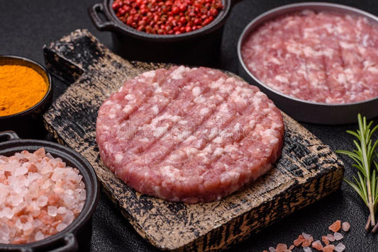 Raw ground pork patty on a stone surface with spices and rosemary in the background