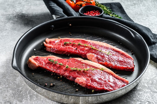 Raw beef steaks on a black grill pan with seasonings on a gray surface