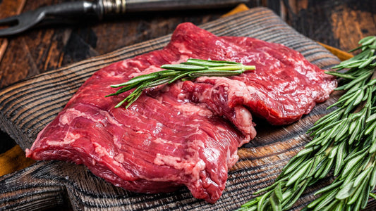 Raw beef skirt on a wooden cutting board with rosemary, surrounded by kitchen utensils.