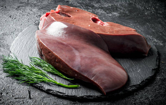 Two pieces of raw liver on a dark slate surface with a sprig of dill.