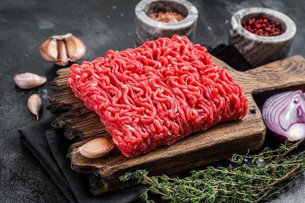 Raw ground meat on a wooden cutting board with garlic, herbs, and spices in the background.