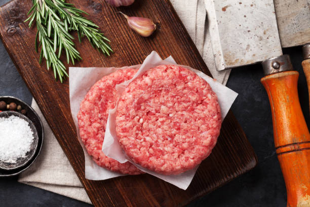 Two raw hamburger patties on a wooden cutting board with rosemary and a cleaver in the background.