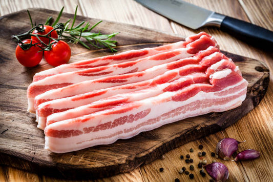 Raw bacon strips on a wooden cutting board with tomatoes and a knife.